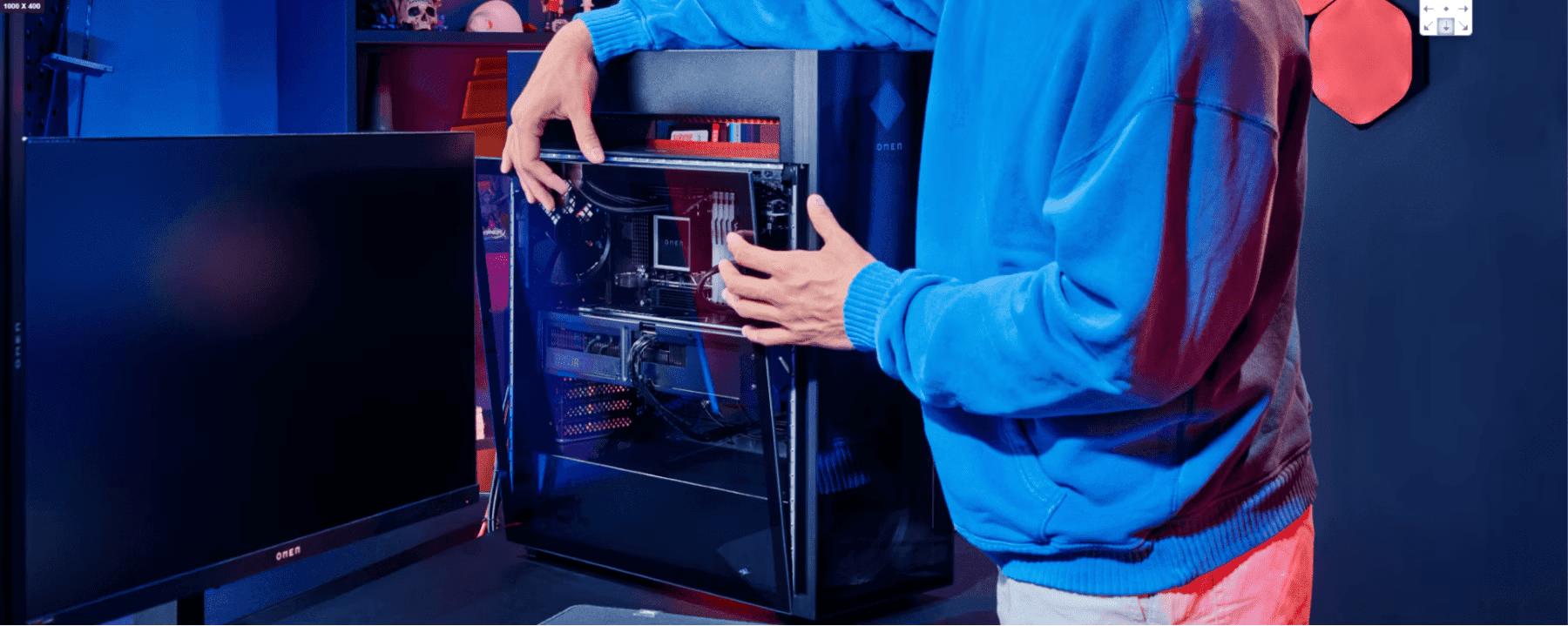 Una persona con sudadera azul sentada frente a una configuración gaming con una caja de PC abierta que muestra componentes internos, iluminada por luces azules y rojas en una habitación oscurecida con decoraciones de pared hexagonales